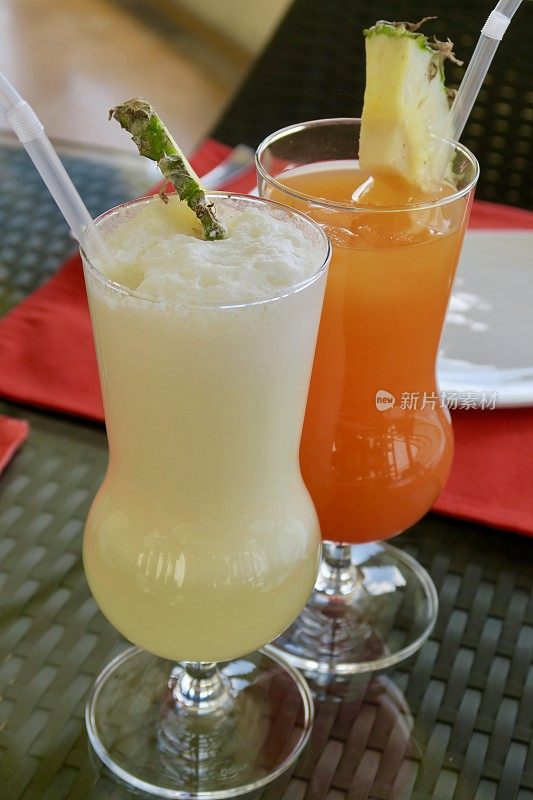 图片:“Piña colada”酒精鸡尾酒/“橙色日落”非酒精鸡尾酒在节日的飓风杯，菠萝装饰和吸管，印度酒吧背景照片